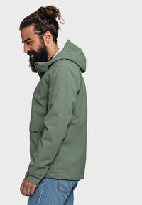 Giacca impermeabile verde con cappuccio, dotata di tasca frontale, chiusura con zip e una texture liscia. Logo visualizzato sulla spalla sinistra.