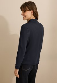 Cardigan texturé bleu marine avec un col haut et des poignets côtelés. Équipé de poches latérales et d'un ourlet droit, associé à un pantalon sombre.