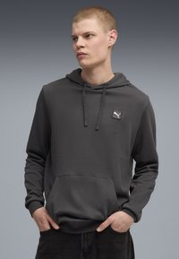 Felpa con cappuccio grigia scura realizzata in morbido tessuto, dotata di una tasca a canguro frontale e un piccolo logo bianco applicato sul petto. Design semplice e moderno.