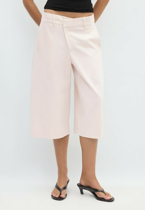 Femme portant un pantalon large court rose pâle et des sandales à talons noires, debout devant un fond clair uni.