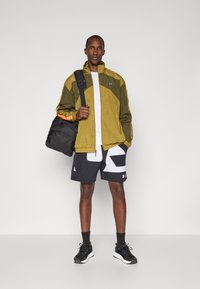 Oliven grøn vindtett jakke med oransje detaljer, hvit t-skjorte, svarte shorts med et markant hvitt mønster, og svarte joggesko med mørke sokker.