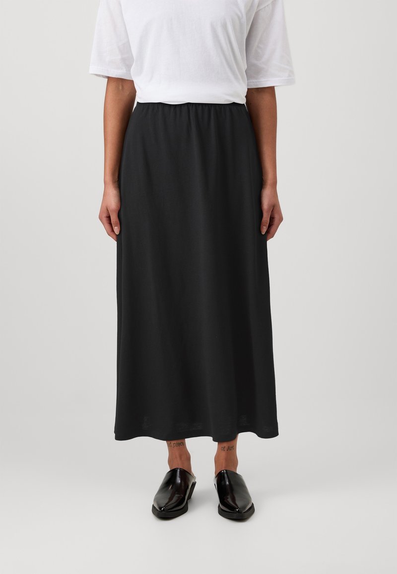 Jupe noire longueur cheville en tissu doux avec une taille élastique. Portée avec une chemise blanche et des chaussures noires sans lacets.