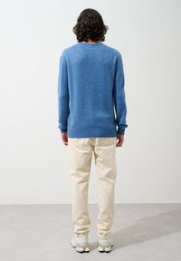 Blå stickad tröja med rund hals, ribbade ärmmuddar och nederkant, kombinerad med beige byxor och vita sneakers, sedd bakifrån.