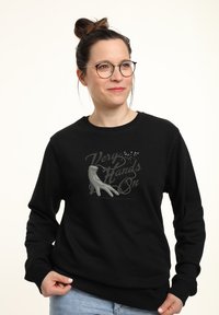 Žena s brýlemi a drdolem, oblečená v černé mikině se šedou rukou a textem "Very Hands On", držící lem u světle modrých džín.