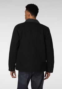 Svart lerretsjakke med en denimkrage, med lange ermer, knappmansjetter og en rett design. Brukes med mørke denimjeans.