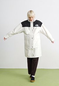 Veste imperméable blanche avec épaules noires, boutons-pression et deux poches à l'avant. Le modèle l'associe à un pantalon marron et des chaussures noires.
