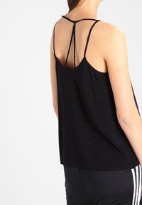 Donna che indossa un top senza maniche nero con sottili spalline doppie che formano un design triangolare sulla parte superiore della schiena, abbinato a pantaloni neri.