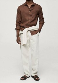 Chemise en lin marron à coupe décontractée, pantalon blanc en tissu tissé, et sandales foncées. Un pull léger est noué autour de la taille.