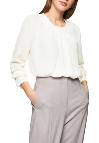 Frau trägt eine langärmelige weiße Bluse, die in eine hoch taillierte hellgraue Hose gesteckt ist, Hände in den Taschen, vor weißem Hintergrund.