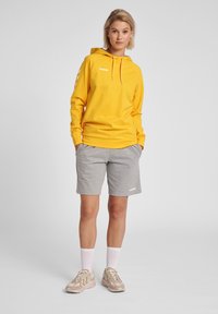 Gul hoodie med dragsko, sidologotyp, kombinerad med grå shorts. Modellen har på sig vita strumpor och beige sportskor med detaljer.