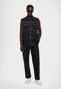 Black sleeveless shirt with two chest pockets and contrasting white stitching, paired with matching trousers featuring patch pockets. Worn with white shoes.