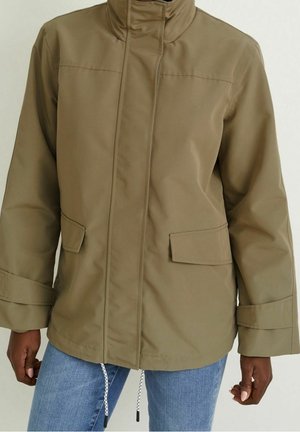 Personne portant une veste marron clair à manches longues avec poches à rabat à l'avant et poignets réglables, associée à un jean bleu.