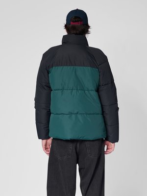 Mężczyzna w czarnej i ciemnozielonej kurtce puchowej oraz czarnych spodniach, zwrócony tyłem, z granatowo-czerwoną czapką baseballową na jednolitym tle.