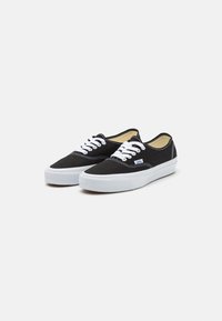 Black canvas sneakers with white laces, white rubber soles, and contrasting white stitching details. Features a branded tag on the side.
