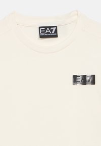 Kreemjas lühikeste varrukatega T-särk, valmistatud pehmest puuvillast, millel on vasakul rinnal musta metallist brändi logoga plaastr. Klassikalise kaelusega disain.