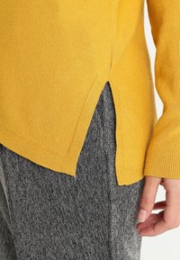 Pull en maille ribbed jaune avec fente sur le côté, associé à un pantalon gris texturé. Le pull présente des manches longues et une coupe décontractée.