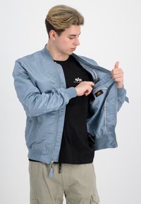 Svetlo modra bomber jakna z zadrgo, rebrastimi manšetami in notranjim žepom. Nosi se čez črn majico in bež hlače.