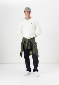 Hvit langermet genser med flerfargede halslinjer, grønn jakke knyttet rundt livet, mørke jeans og hvite joggesko.