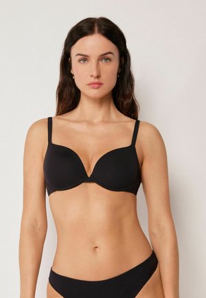 Femme aux longs cheveux noirs portant un soutien-gorge noir et une culotte assortie, debout contre un fond clair uni.