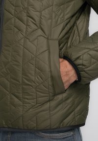 Grüne, gesteppten Jacke mit einem strukturierten Muster, schwarzem Besatz und einer Seitentasche, in die eine Hand eingesteckt ist. Das Material scheint synthetisch und leicht zu sein.