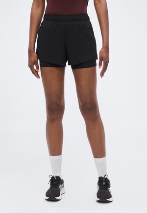 Personne portant un short de sport noir superposé, des chaussettes blanches montantes et des chaussures de course noires, debout sur un fond clair.