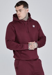 Felpa bordeaux con tasca frontale, cappuccio con coulisse e un piccolo logo bianco sul petto. Tessuto morbido con una vestibilità comoda.