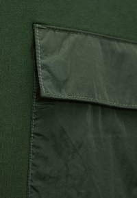 Primer plano de una tela verde oscura con un bolsillo rectangular bordado que presenta una solapa sobre un fondo texturizado.