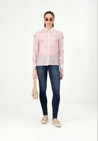 Camisa transparente rosa clara com detalhes em fita rosa, jeans skinny azuis, sapatos rasos beges e bolsa a condizer. Óculos de sol redondos rosa.