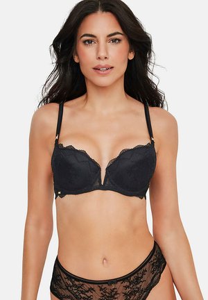 Femme aux longs cheveux foncés portant un soutien-gorge en dentelle noire et une culotte assortie en dentelle noire, faisant face à l'avant sur un fond uni.