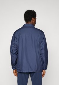 Man wearing a navy blue lightweight jacket and matching pants, standing facing away against a plain light background.