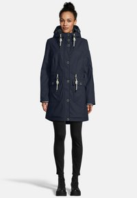 Vrouw in een marineblauwe gewatteerde parka met witte trekkoorden, zwarte leggings en zwarte veterschoenen, staand op een witte achtergrond.