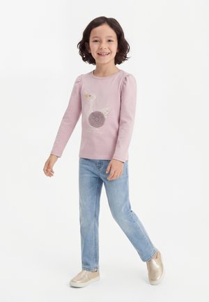 Uśmiechnięte dziecko idące, ubrane w jasnofioletową bluzkę z długim rękawem z wzorem łabędzia, niebieskie dżinsy i błyszczące beżowe buty wsuwane na białym tle.