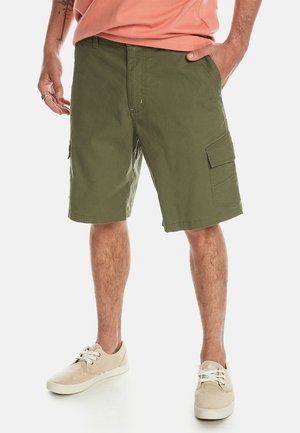 Cargo-Shorts aus olivgrünem Baumwollstoff mit Seitentaschen, einem lockeren Schnitt und einem Saum, der oberhalb des Knies endet. Getragen mit beigen Schuhen.