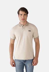 Polo beige in cotone con colletto e polsini a coste, caratterizzato da un piccolo badge nero sul petto. Indossato con jeans blu.
