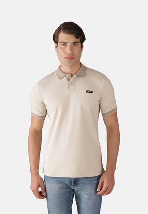 Polo beige in cotone con colletto e polsini a coste, caratterizzato da un piccolo badge nero sul petto. Indossato con jeans blu.