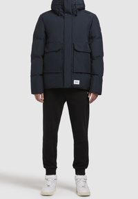 Granatowa pikowana kurtka z wysokim kołnierzem, kapturem ze ściągaczem, dwoma przednimi kieszeniami oraz naszywką z logo na dole. Do tego czarne spodnie dresowe i białe trampki.