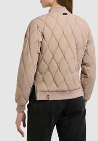 Femme portant une veste matelassée marron clair avec des poignets et un ourlet côtelés, associée à un jean en denim noir, vue de dos.