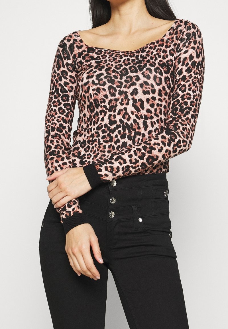 Femme portant un haut manches longues à imprimé léopard rose et noir, associé à un pantalon noir taille haute avec des boutons argentés, les bras croisés.