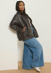 Giacca di pelle marrone con un design colletto, dotata di tasche laterali e polsini a coste. Abbinata a jeans in denim blu a gamba larga.