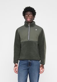 Fleece pullover in dark green with a high collar, half-zip, and textured body. Accented with a smooth fabric on sleeves and logo detail.