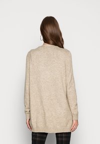 Donna con lunghi capelli castani che indossa un maglione di maglia beige e pantaloni a quadri neri, in piedi di fronte a uno sfondo bianco semplice.