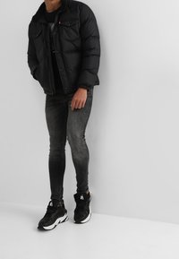 Chaqueta acolchada negra con botones a presión, camiseta gráfica gris debajo, jeans skinny negros desgastados y zapatillas negras con acentos blancos.