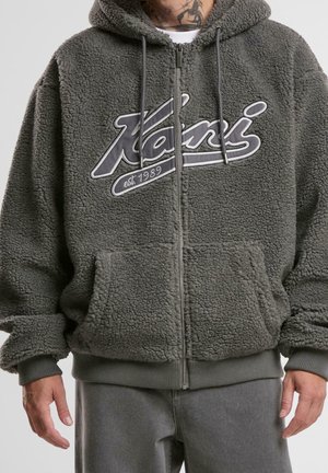 Mand iført grå, fluffy lynlåstrøje med forlommer, "Kassi est. 1989" tekst på brystet og synlig hals tatovering med skorpion.