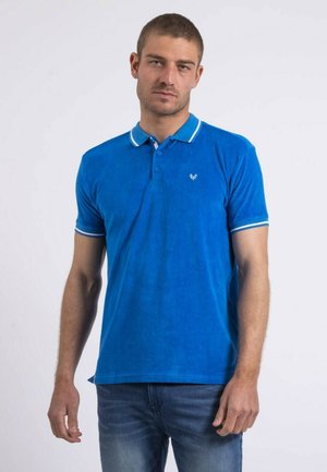 Homme portant un polo à manches courtes bleu vif avec un bord blanc sur le col et les manches, debout devant un fond clair uni.