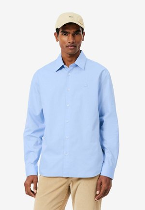 Hombre con gorra beige, camisa azul claro abotonada y pantalones de pana beige, de pie frente a un fondo blanco liso.