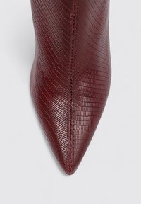 Botín burdeos de punta afilada con un material texturizado inspirado en reptiles. Presenta una silueta elegante y una costura que recorre la parte frontal.