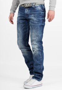 Jean en denim bleu moyen avec un lavage délavé, coupe slim et coutures visibles. Associez-les à des baskets blanches avec des accents rouges.