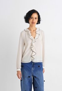 Cream blouse with a ruffled neckline, black piping, and long sleeves, paired with blue denim trousers featuring black pom-pom accents.
