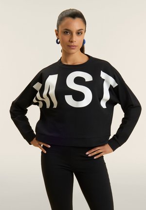 Nainen, jolla on tummat hiukset poninhännällä, musta collegepaita, jossa isot valkoiset kirjaimet "M S T", ja mustat housut, kädet lanteilla.