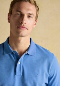 Sinine puuvillane polo särk, millel on kahe nupuga kaelus, lühikesed varrukad ja rindkere peal väike broderi logo. Sile tekstuur ja juhuslik disain.